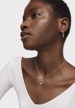 Collana d'argento con un pendente astratto unico che presenta una perla, indossata su una maglietta bianca a scollatura a V. Primo piano su scollatura e orecchini.