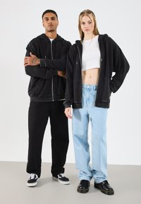 Čierne mikiny na zips s prednými vreckami, nosené cez biely crop top; skombinované s čiernymi a svetlo modrými širokými nohavicami a čiernymi topánkami.