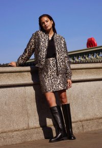 Béžová a černá látková bomber bunda s leopardím vzorem a sukně, doplněná černými koženými kolenními botami. Venku, za jasného nebe.