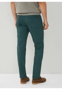 Man in een teal slim-fit broek en bruine suède schoenen, staand tegen een effen lichtgrijze achtergrond, gezien van achteren.