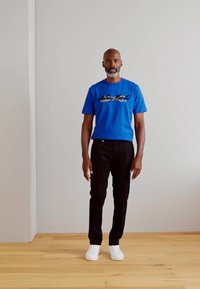 T-shirt in cotone blu con scritte nere, abbinata a pantaloni neri e sneaker bianche. Il modello si trova davanti a uno sfondo neutro.