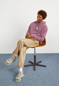 Sudadera lila claro con un logo en el frente, pantalones beige y zapatillas grises con cordones multicolores. Sentado en una silla marrón frente a un fondo neutro.