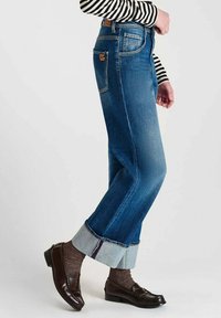 Jean en denim bleu clair avec un design large et cropped. Comprend un ourlet replié, cinq poches et une étiquette en cuir marron à l'arrière.