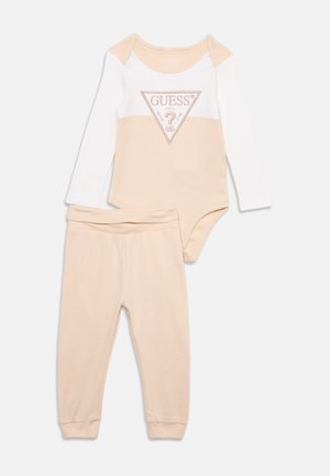 Ensemble bébé deux pièces avec pantalon beige et body manches longues beige et blanc avec logo Guess sur la poitrine.