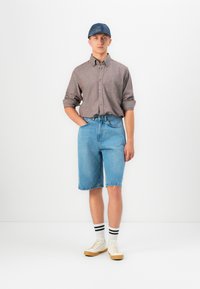 Joven de pie con la mano en el bolsillo, vistiendo una gorra azul, una camisa marrón de botones, pantalones cortos de mezclilla, calcetines blancos con rayas negras y zapatillas beige.