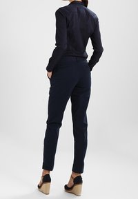 Tmavě modrá upnutá košile s límečkem, dlouhými rukávy a detailem knoflíků, spárovaná s tmavými kalhotami a sandály na platformě s tkaným designem.