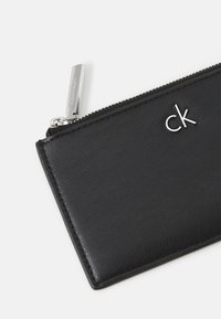 Svart lær lommebok med en glatt tekstur, med en sølvfarget glidelås og en liten metallisk "ck"-logo på forsiden. Kompakt rektangulær form.