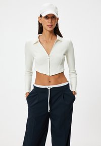 Weißes, geripptes Crop-Top mit Reißverschluss vorne, mit Kragen und langen Ärmeln. Kombiniert mit dunkelblauen Hosen mit Kordelzugbund.