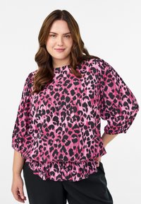 Ženska z dolgimi rjavimi lasmi oblečena v bluzo v roza in črnem leopardjem vzorcu z nabranimi rokavi in črnimi hlačami, stoji pred belo ozadjem.