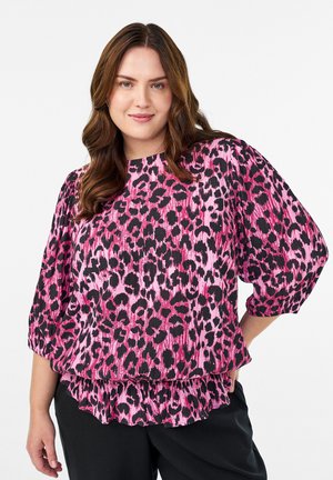 Kvinde med langt brunt hår iført en bluse i pink og sort leopardprint med pufærmer og sorte bukser, stående mod en hvid baggrund.