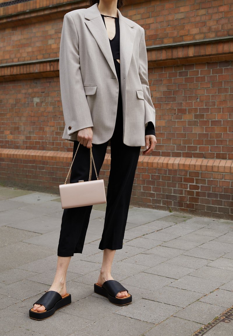 Blazer beige overdimensionato con due tasche frontali, top corto nero sotto, pantaloni larghi neri, sandali platform neri e una borsa a tracolla rosa.