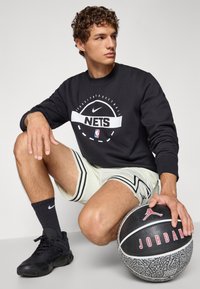 Sort sweatshirt med "NETS" logo, lyse perforerede shorts med striber, sorte sneakers, holder en basketball med "JORDAN" mærkning.
