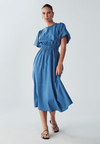 Modra midi obleka z okroglim izrezom, napihnjenimi rokavi in nabranim pasom. Narejena iz mehkega materiala; zračen, gladek tekstura, v kombinaciji z črnimi sandali.