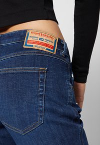 Jeans de ganga azul com um rótulo de couro visível apresentando "Diesel Industry", costura laranja e design padrão de bolso traseiro.