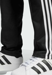 Czarne spodnie sportowe z trzema pionowymi białymi paskami i białe trampki z czarnymi paskami oraz teksturowanym noskiem.