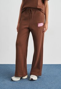 Καφέ παντελόνια τζόγκερ κατασκευασμένα από μαλακό ύφασμα, με ευρύ σχεδιασμό ποδιού, που διαθέτουν ροζ κεντημένο λογότυπο Nike στο αριστερό μηρό. Στυλιζαρισμένα με αθλητικά παπούτσια.