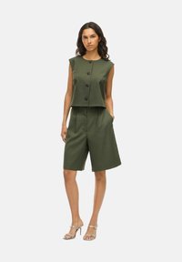 Top senza maniche verde olivo con bottoni frontali, abbinato a pantaloni shorts su misura. Sandali con tacco alto argentati completano il look.