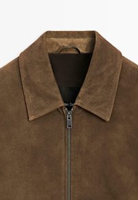 Brun ruskindsjakke med en skarp krave og fuld lynlås foran. Glat tekstur, minimal detaljer og et klassisk design.