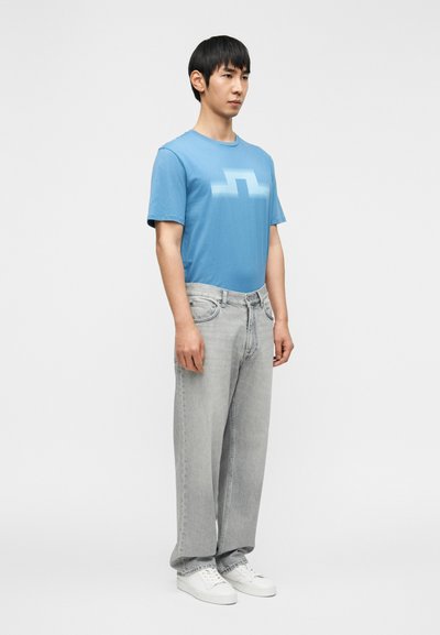 Άνδρας που φοράει μπλε μπλουζάκι με σχέδιο, ανοιχτό γκρι τζιν με φαρδύ πόδι και λευκά αθλητικά, στέκεται μπροστά σε απλό ανοιχτό φόντο.