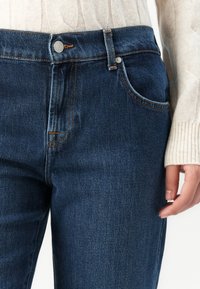 Gros plan sur une personne portant un jean en denim bleu foncé et un pull en laine de couleur crème, avec une main visible sur le côté.