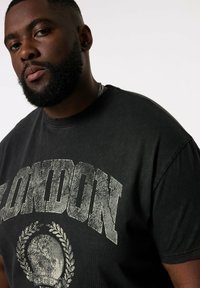 Černé grafické tričko s krátkým rukávem a kulatým výstřihem, které má opotřebovaný tisk "LONDON" a kruhovou grafiku s vavřínem.