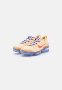 Nike Air VaporMax-skor i ljusbeige med lila snören. Har en strukturerad ovandel och transparent gummisula, accentuerad av en röd logotyp.