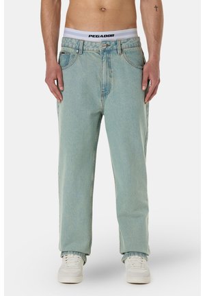 Homme portant un jean bleu clair ample, des baskets blanches et un sous-vêtement avec ceinture blanche visible avec le texte "PEGADOR".