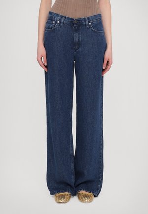 Personne portant un jean denim bleu foncé à jambes larges, un haut en maille marron et des chaussures plates tissées dorées, debout devant un fond clair uni.