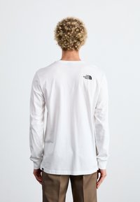 Langärmliges weißes T-Shirt mit rundem Ausschnitt, das ein schwarzes Logo auf dem oberen Rücken trägt. Aus weicher Baumwolle mit glatter Textur hergestellt.