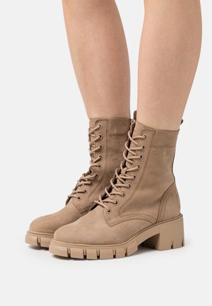 Bottes de combat en daim beige avec bout rond, lacets sur le devant et semelle en caoutchouc épaisse. Présentent des détails de couture et une finition texturée.