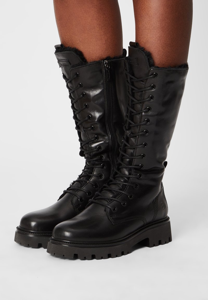 Botas de cuero negro hasta la rodilla con un acabado brillante, cordones en la parte delantera, cremallera lateral y ribete de piel de textura en la parte superior. Suela de goma gruesa.