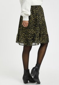 Jupe à imprimé léopard en vert et noir, avec un ourlet à volants. Portée avec des bottines noires et des collants opaques. Tissu texturé.