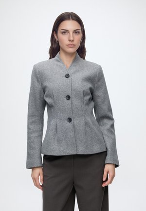 Femme aux cheveux foncés portant une veste grise cintrée boutonnée et un pantalon sombre, debout devant un fond clair uni.
