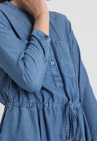 Robe en denim à manches longues, avec boutonnière sur le devant, une poche poitrine unique, taille élastique et détail noué, de couleur bleu clair et texture douce.