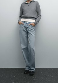 Harmaa pitkähihainen polo, vaaleanvärinen aluspaita, vaaleansiniset suorat farkut ja mustat jalkineet neutraalia taustaa vasten.