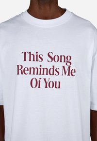 Valge puuvillane T-särk, millel on tekst "See laul meenutab mind sinust" burgundi värviga, tsentriteeritud julge ja modernse fondiga. Lühikesed varrukad.