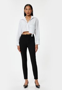 Camisa blanca corta con un bolsillo en el pecho y un detalle de corbata. Pantalones negros ajustados con un diseño clásico. La modelo lleva unos zapatos planos negros con punta.