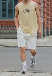 Beige mouwloos sporttop met zwart logo, witte shorts en grijze sneakers. Inclusief een zilveren ketting en witte sokken. Urban omgeving.