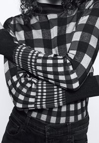 Pull à carreaux noir et blanc avec manches côtelées noires, présentant des carreaux de tailles variées et un col montant. Matière tricotée texturée.