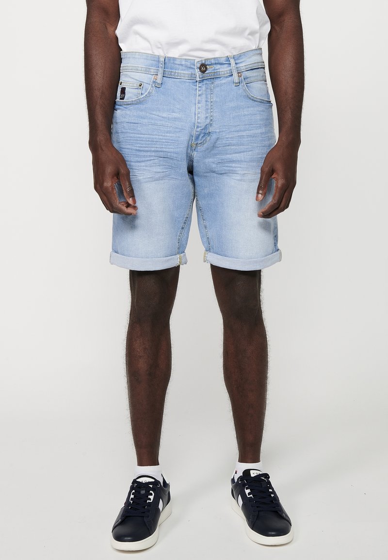 Homme portant un short en jean bleu clair retroussé, des chaussettes blanches, des baskets bleu marine et une chemise blanche, debout devant un fond uni.