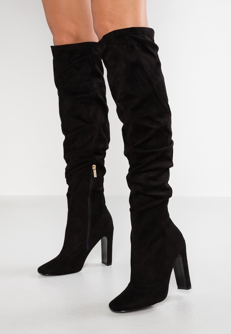 Botas de ante negras por encima de la rodilla con un diseño ajustado, punta afilada y un tacón alto y en bloque. Cuenta con un cierre de cremallera lateral para facilitar su uso.
