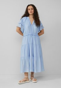 Helles blaues, gestuftes Kleid mit Rüschensleeves und gerafftem Taillenbereich. Hergestellt aus einem weichen Stoff mit fließender Silhouette. Getragen mit Riemchensandalen.