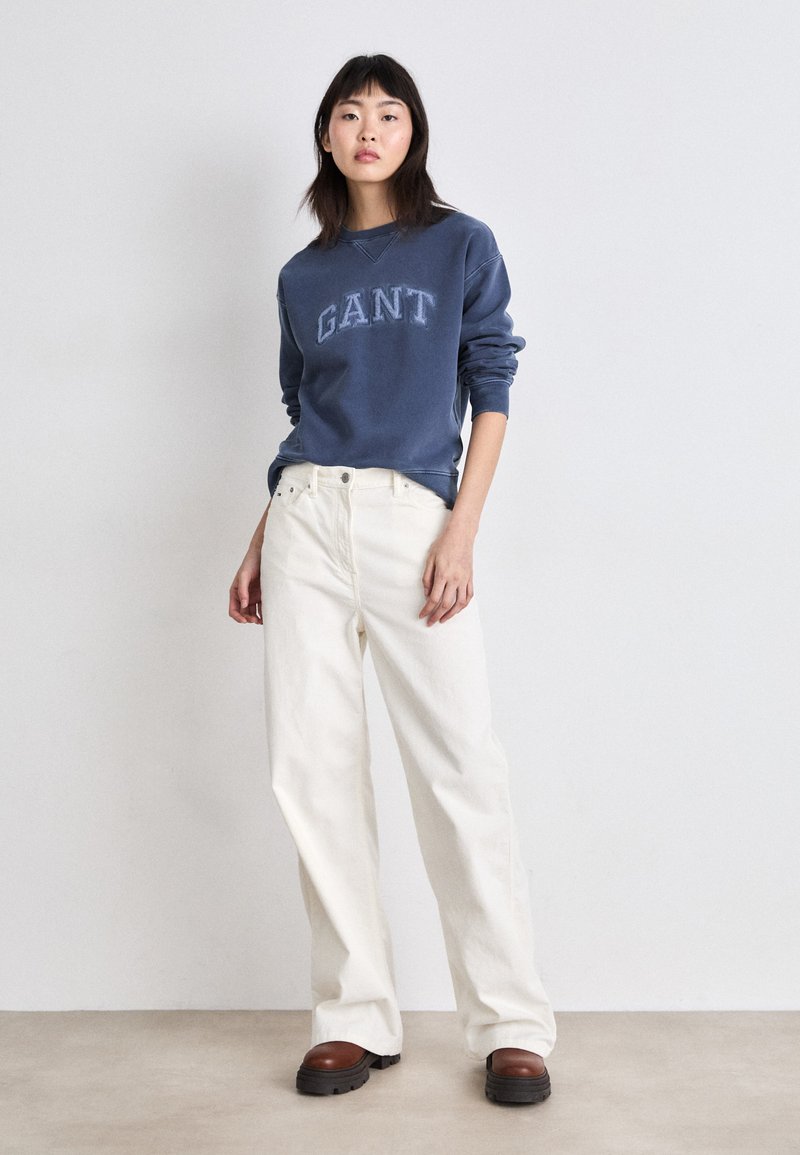 Granatowa bluza z logo "GANT", zestawiona z białymi szerokimi spodniami dżinsowymi i brązowymi butami na platformie, charakteryzująca się luźnym krojem i miękką fakturą.