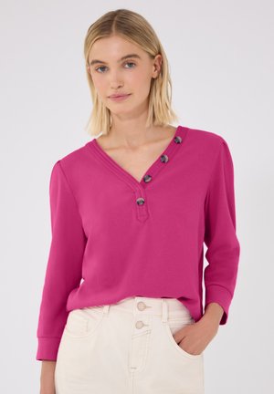 Jonge vrouw met kort blond haar die een roze top met lange mouwen draagt met vier knopen bij de V-hals en een lichte broek.