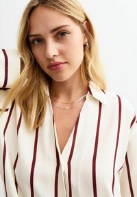 Witte blouse met verticale bordeauxstrepen, met een kraag en een V-hals. Het model draagt gelaagde zilveren kettingen en heeft golvend blond haar.