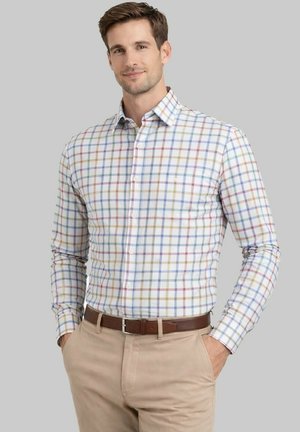 Homme portant une chemise à manches longues blanche avec un motif à carreaux multicolores, un pantalon beige et une ceinture marron, debout les mains dans les poches sur un fond gris.