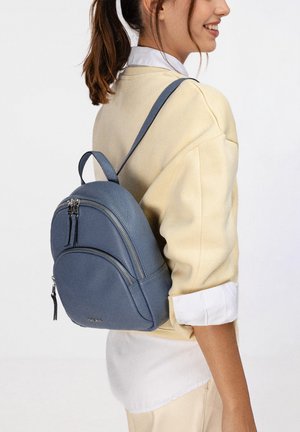 Jeune femme portant un pull beige et une chemise blanche, elle porte un petit sac à dos en cuir bleu avec deux compartiments zippés.