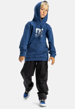 Blauwe hoodie met een voorzak en wit logo, gecombineerd met zwarte broek en blauwe sneakers met witte en zwarte accenten.