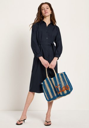 Frau trägt ein schwarzes Hemdblusenkleid mit Gürtel und schwarze Sandalen mit Absatz, hält eine blau-beige gewebte Tragetasche mit orangefarbener Schrift.