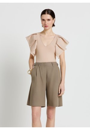 Donna con top beige con maniche a volant stratificate e pantaloncini a vita alta color verde oliva di lunghezza fino al ginocchio con tasche, in piedi contro uno sfondo bianco.